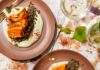 Roasted Butternut Squash With Lentils and Whipped Feta Roasted butternut squash wedges served over green lentils and whipped cilantro feta, garnished with crushed pistachios and Aleppo pepper, paired with white wine glasses.