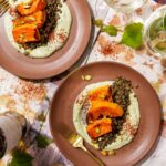 Roasted butternut squash wedges served over green lentils and whipped cilantro feta, garnished with crushed pistachios and Aleppo pepper, paired with white wine glasses.