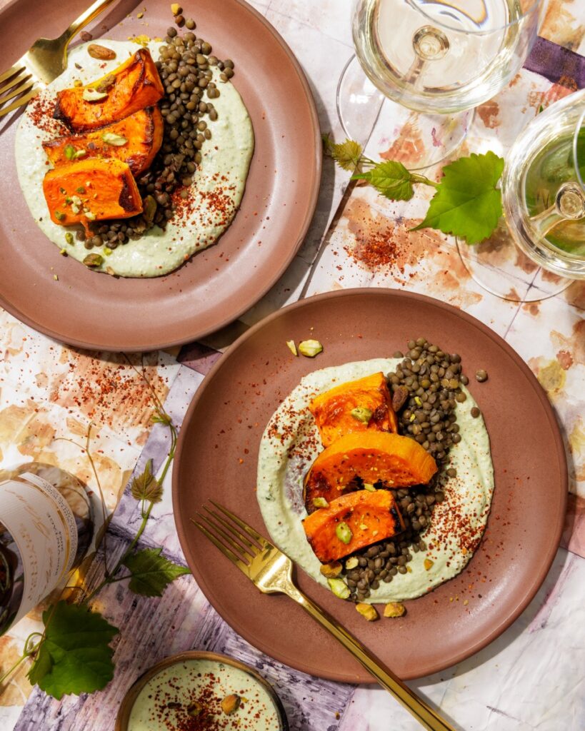 Roasted butternut squash wedges served over green lentils and whipped cilantro feta, garnished with crushed pistachios and Aleppo pepper, paired with white wine glasses.