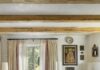 Designing a Small Santa Fe Condo to Feel Complete Living room centered around a rustic wood trunk coffee table, blue sofa, two leather armchairs, a white kiva-style fireplace, exposed ceiling vigas, and double-window treatments with cream drapes and sheer panels.