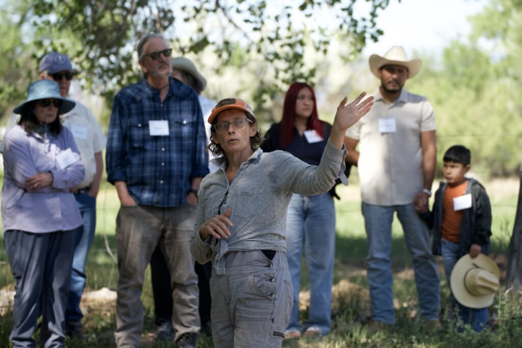 A female farmer wearing a cap, glasses, and overalls gesturing with her hand while leading a group tour outdoors in a field.