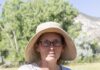 Faces of the Santa Fe Farmers’ Market: The Story of Malandro Farm Portrait of a female farmer wearing glasses and a sunhat, leaning against a fence outdoors with a field and mountains in the background.