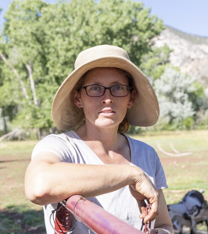 Portrait of a female farmer wearing glasses and a sunhat, leaning against a fence outdoors with a field and mountains in the background.