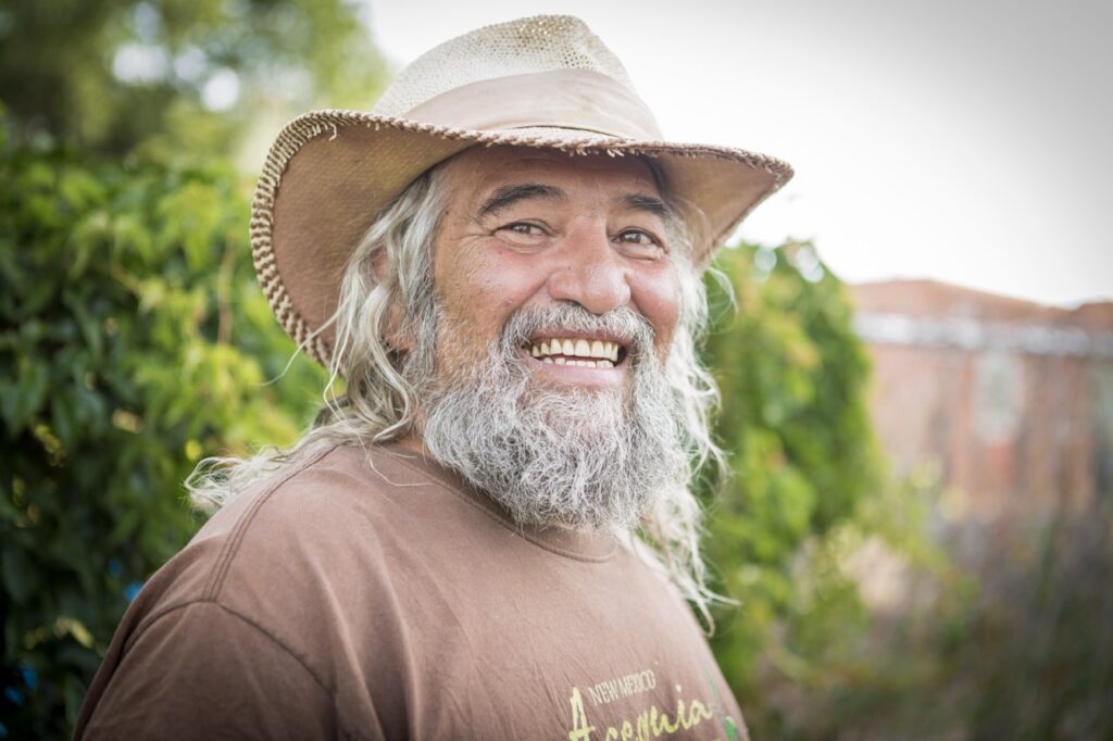 A man in a brown hat with a grey beard and hair and brown shirt.