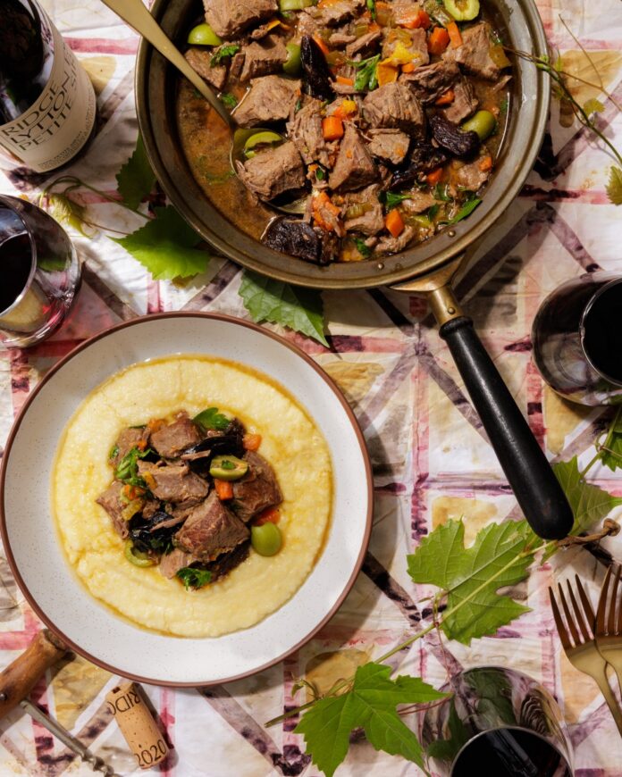 Slow-cooked beef stew braised with prunes, green Castellano olives, and orange peel, served over smooth polenta alongside glasses of red wine and a bottle of Ridge Lytton Estate Petite Sirah.
