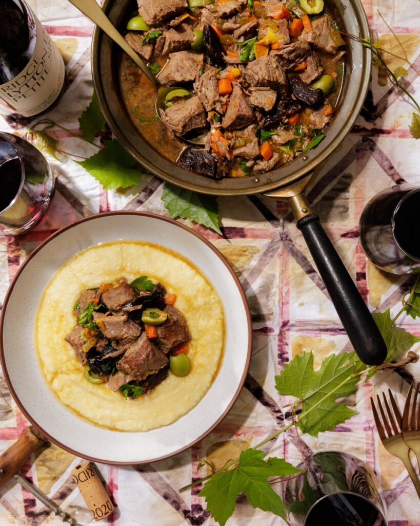 Slow-cooked beef stew braised with prunes, green Castellano olives, and orange peel, served over smooth polenta alongside glasses of red wine and a bottle of Ridge Lytton Estate Petite Sirah.