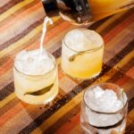The 505 mezcal cocktail being poured from a shaker into rocks glasses over crushed ice, garnished with burnt sage leaves on a striped woven placemat.
