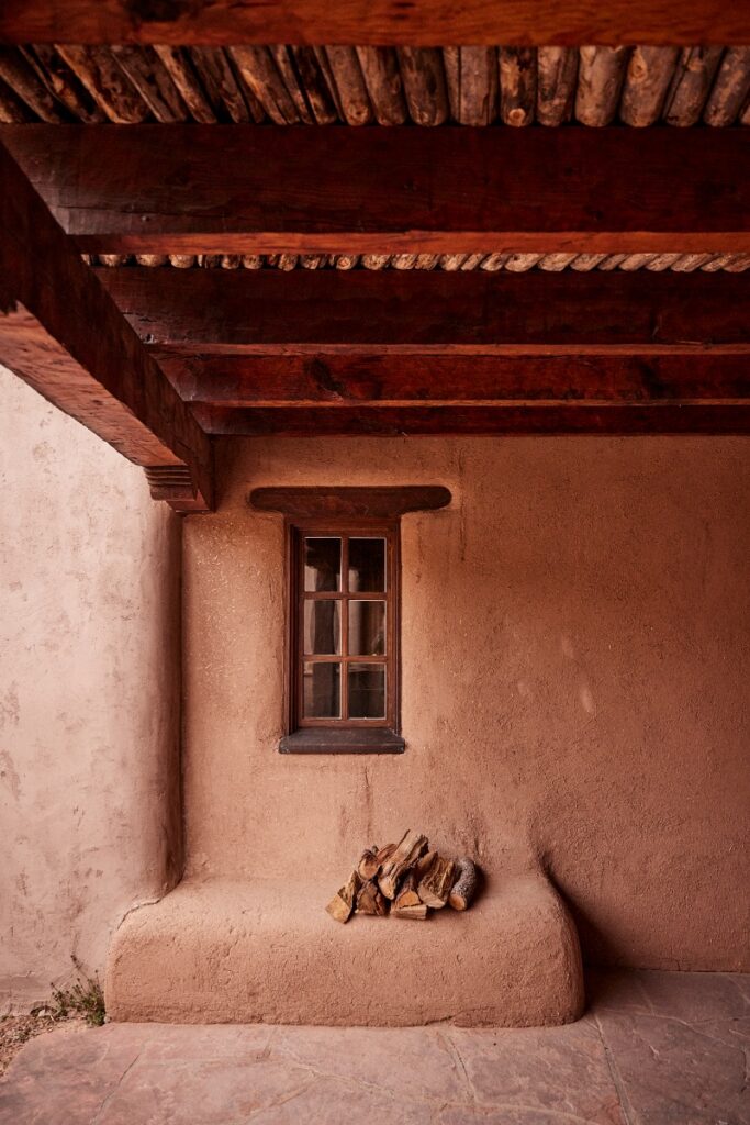Exterior detail of the historic adobe home featuring thick earthen plaster walls, a small multi-pane window, and exposed wood beam ceilings over a bench with stacked firewood.