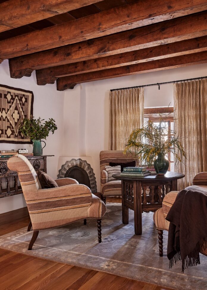 Warm living space styled by French & French, featuring heavy dark wood ceiling beams, striped armchair seating, neutral curtains, a Native American wall hanging, and a small kiva fireplace.