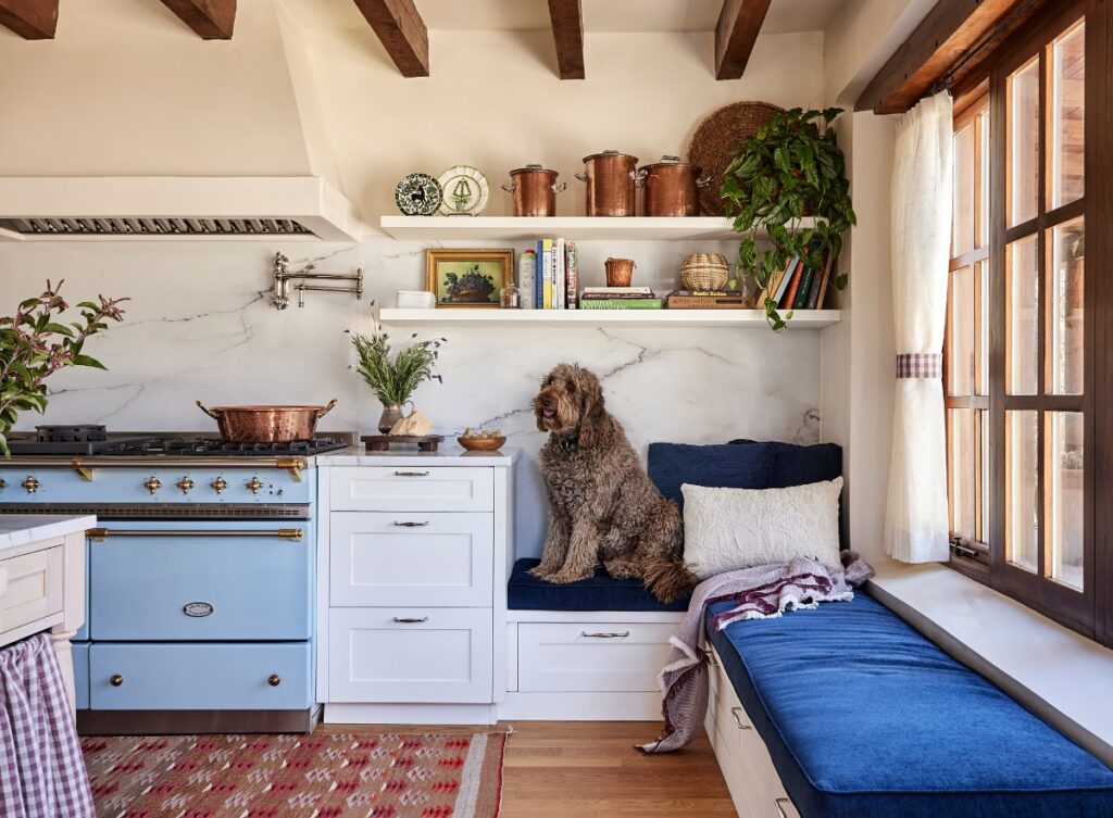 Bright kitchen area featuring a light blue range stove, marble backsplash, open shelving, and a dark blue upholstered window seat with a dog lounging on top.