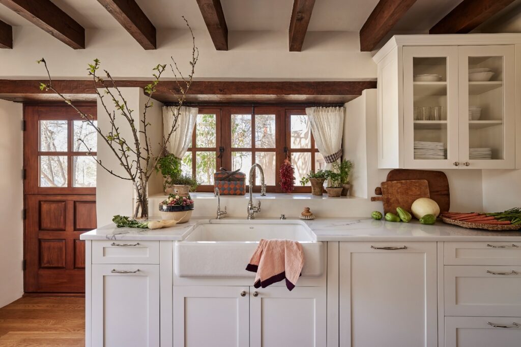 Sunlit kitchen apron-front sink positioned beneath wood-framed grid windows, featuring white cabinetry, marble countertops, and rustic wooden ceiling beams.