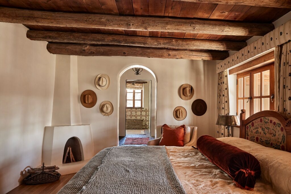 Cozy primary bedroom with a traditional corner kiva fireplace, dark timber ceiling beams, woven textiles, and wide-brimmed hats mounted on the white plaster wall.