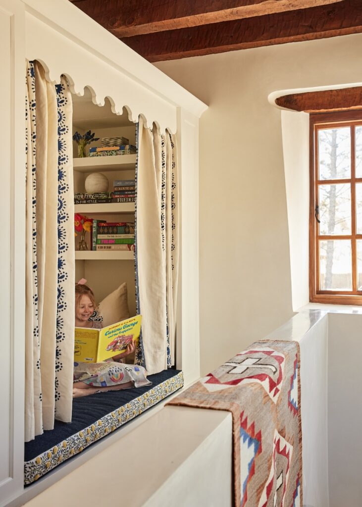 A custom built-in reading nook set inside an adobe wall alcove with blue-and-white patterned curtains, where a young child reads a book.