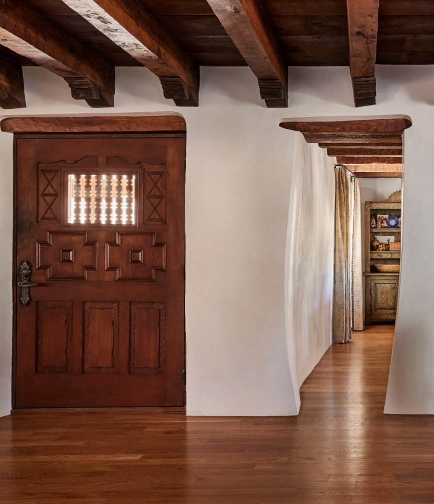 Interior hallway view showing an intricately carved dark wood door alongside smooth, thick plastered white adobe walls leading to an adjacent room.