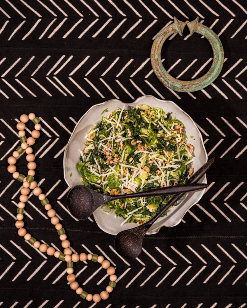 A top-down view of a scalloped ceramic bowl filled with Simple Brussels Sprouts Slaw, set with dark wooden servers on a geometric black-and-white mudcloth textile alongside wooden beads and a green patinated metal ring artifact.
