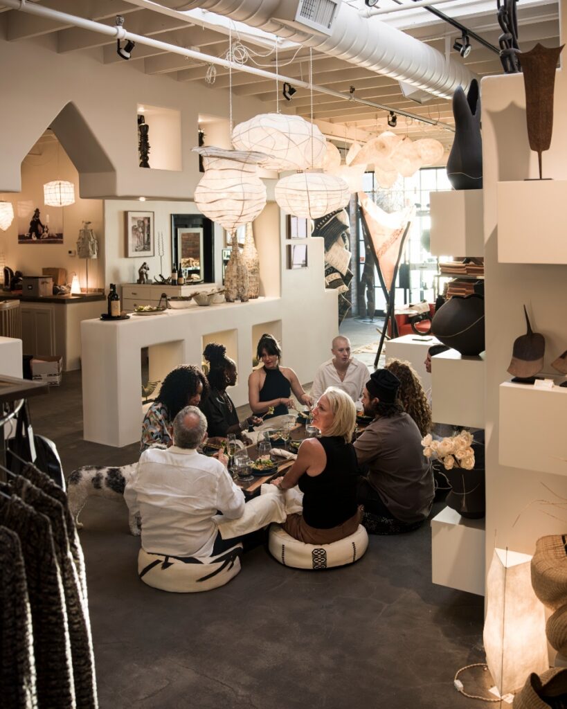 High-angle shot showing the full dinner gathering seated on poufs around a low table under delicate paper pendant lights, with Vanna the dog visible nearby.