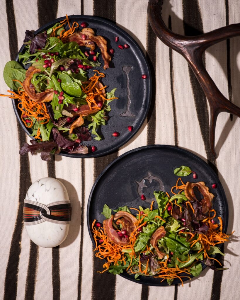 A top-down view of two dark ceramic plates featuring the "I Yam What I Yam" salad topped with greens, pancetta shards, maple-glazed shoestrings, and pomegranate seeds, displayed on a striped fabric next to an animal horn sculpture.