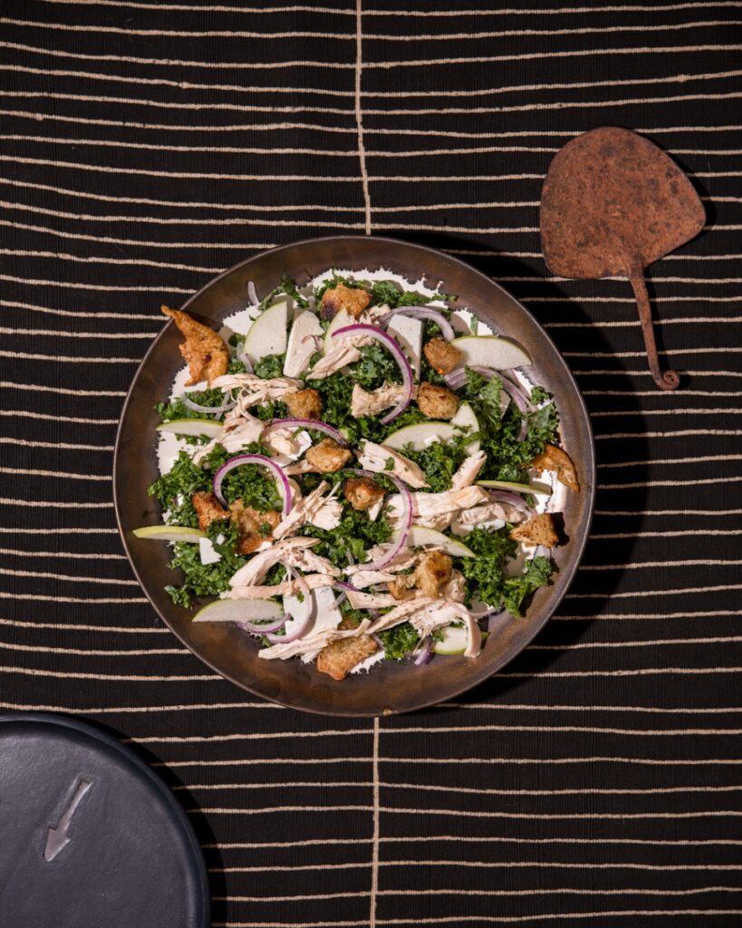 An overhead shot of "The Harvest Moon" chicken salad plated with kale, sliced apples, red onions, croutons, and crackling, placed on a dark striped mat beside a weathered metal tool.