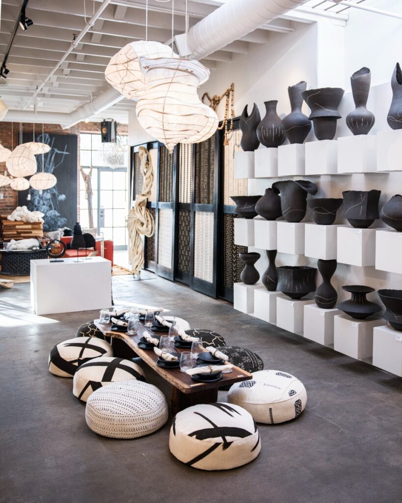 Wide interior view of the Vanna gallery displaying paper lantern pendant lights, floor poufs around a low wooden dining table, and shelving stacked with organic black ceramic vessels.