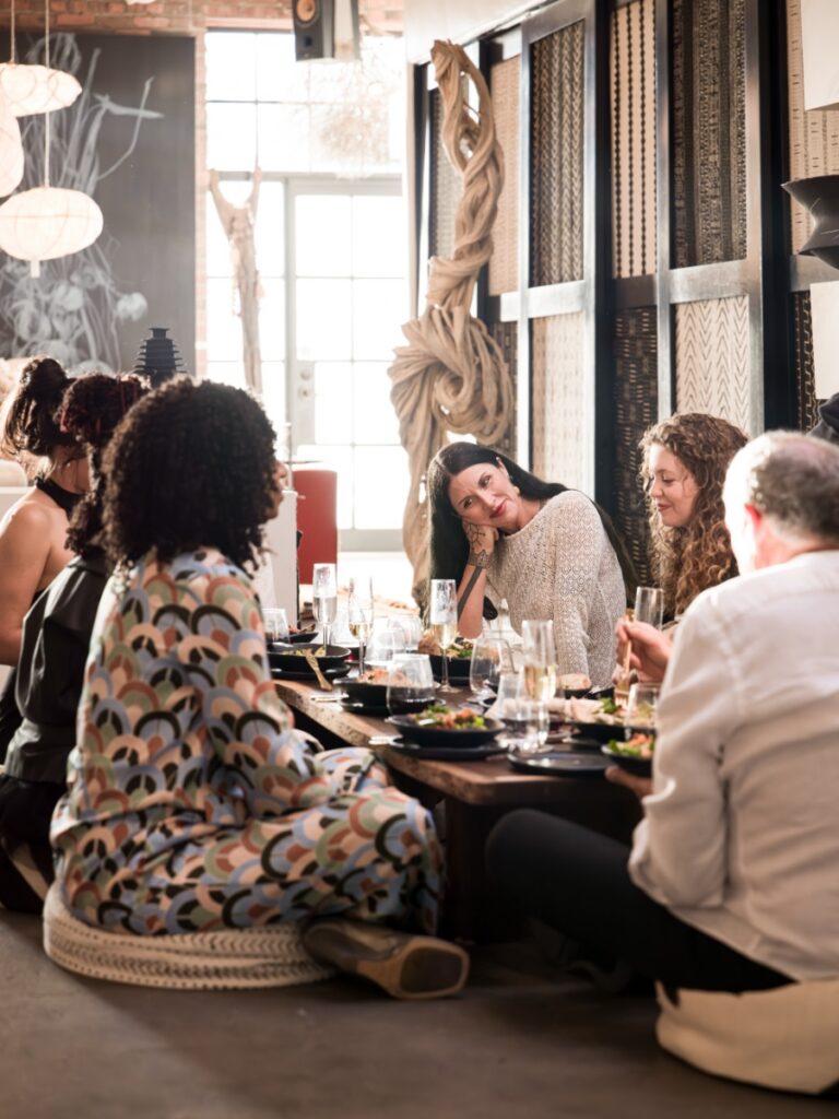 Dinner guests seated around a low wooden table on floor poufs, conversing and sharing a meal within the warmly lit gallery environment.