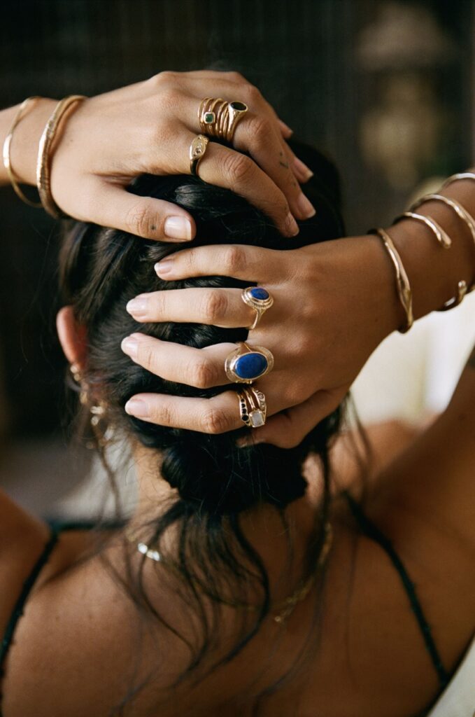 A woman holds the back of her head with many different rings and bangles on her hands and wrists.