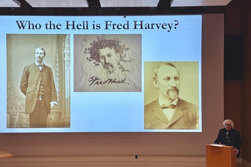 A man at a podium gives a presentation on Fred Harvey with a slide presenting "Who the Hell is Fred Harvey?"