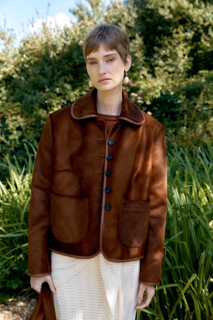 A woman with short brown hair wearing a brown bomber jacket and white skirt outside.